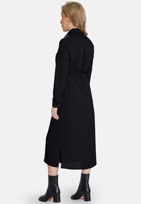 Abito camicia nero a maniche lunghe realizzato in tessuto morbido, con vita aderente, spacchi laterali e colletto. Indossato con stivali neri alla caviglia.