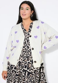 Kremowy sweter na guziki z fioletowymi wzorami serc, o luźnym kroju, w zestawieniu z czarną i beżową sukienką w panterkę.