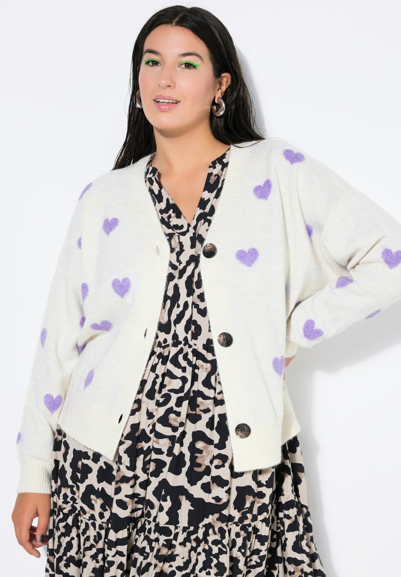 Kremowy sweter na guziki z fioletowymi wzorami serc, o luźnym kroju, w zestawieniu z czarną i beżową sukienką w panterkę.