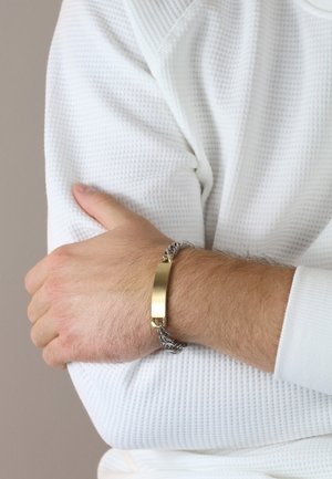 Silberne Kettenarmband mit einem rechteckigen goldenen Mittelpunkt. Das Material ist metallisch mit einer glatten Oberfläche und wird an einem Handgelenk mit strukturiertem Stoff getragen.