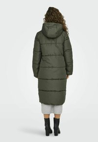 Lange groene pufferjas met een capuchon, voorzien van horizontaal gestikte delen. Gedragen over een grijze rok en zwarte hakken.