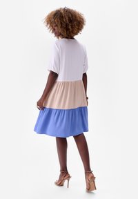 Kleurgebroken jurk met een witte top, een beige middenstuk en een blauwe rok. Korte pofmouwen, ronde halslijn en vloeiende stof. Hakken zichtbaar.