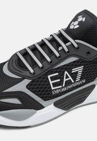 Musta ja harmaa urheilukenkä, jossa on verkkopäällinen, kumipohja ja heijastavat yksityiskohdat. Varustettu säädettävillä nauhoilla ja logotiedolla sivussa.