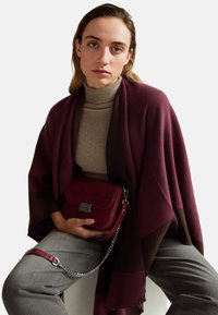 Junge Frau trägt einen beigefarbenen Rollkragenpullover, einen scharlachroten und braunen Schal, graue Hosen und hält eine scharlachrote Handtasche aus Leder mit einer silbernen Kette, während sie sitzt.