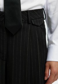 Črno prugaste hlače z teksturiranim zaključkom so opremljene s skrito zapirala, sprednjimi žepi in usklajeno temno kravato s herringbone vzorcem.