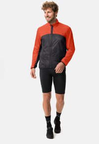 Giacca a vento leggera, con la parte superiore rossa e la parte inferiore nera, dotata di una zip completa e polsini elasticizzati. Indossata con pantaloni corti neri.