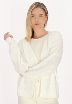 Cardigan bianco e soffice con un design ampio e aperto, dotato di maniche larghe e orlo a coste, indossato sopra una maglietta beige chiaro.