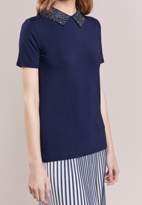 Camisa de manga corta de color marino con un cuello estampado y adornado. Tejido suave, diseño ajustado y dobladillo recto. Combinada con una falda a rayas.