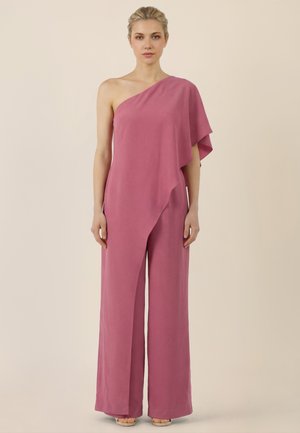 Kvinde iført en ærmeløs, pink jumpsuit med ét skulderstrop og bukser med vide ben, stående foran en ensfarvet beige baggrund.