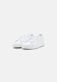 Zapatillas deportivas blancas con un upper sintético liso, punta redonda, cierre con cordones y un logotipo sutil en los laterales. Suela de goma para tracción.