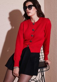 Rote, langärmlige Strickjacke mit sechs schwarz gefärbten Knöpfen in Rosettenform, kombiniert mit einem schwarzen Rock. Glatte Textur und taillierte Passform, sitzend auf einem Drahtstuhl.