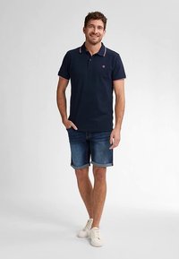 Námořnické polo tričko s kontrastními detaily růžového límce, v kombinaci s džínovými šortkami. Modelka nosí bílé tenisky a pózuje nenuceně s rukama v bok.