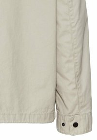 Lys beige jakke med en glat tekstur. Har en knap ved manchetterne og en lige nederdel med minimal syning.