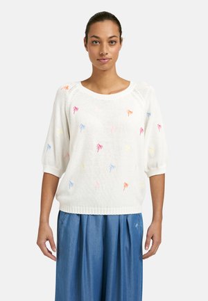 Frau trägt einen weißen Pullover mit bunten Palmenmustern und blaue weit geschnittene Hosen, steht vor einem schlichten hellen Hintergrund.