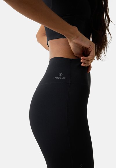 Frau, die hoch taillierte schwarze Leggings mit dem Logo "Fire+Ice" anpasst, seitlichen Torso und Hand vor weißem Hintergrund zeigend.
