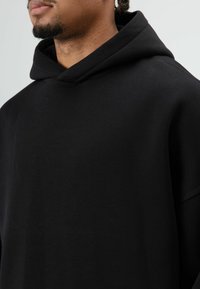Zwarte hoodie gemaakt van zachte stof, met een losse pasvorm, wijde mouwen en een hoge halslijn. Eenvoudig ontwerp zonder zichtbare patronen of accenten.