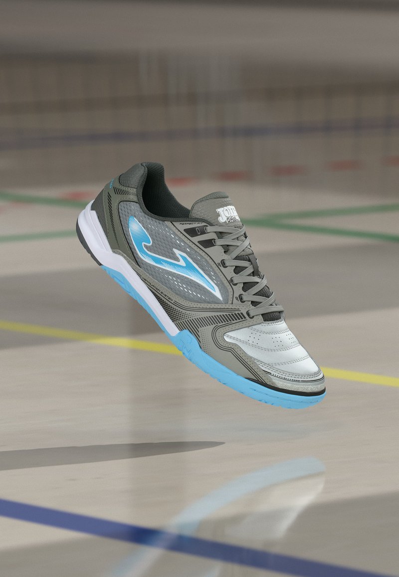 Chaussure de sport d'intérieur grise et bleue suspendue au-dessus d'un sol de gymnase avec des lignes de terrain visibles en dessous.