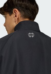 Chaqueta negra con cuello alto y un pequeño emblema blanco en la parte trasera. Textura suave con un brillo sutil y costuras planas.