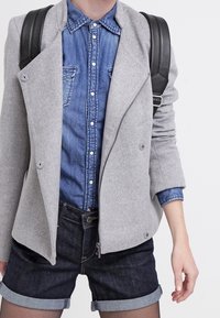 Chaqueta gris de mezcla de lana de doble botonadura sobre una camisa de mezclilla de botones, combinada con unos pantalones cortos de mezclilla oscuros arremangados. Se ven las correas de una mochila negra.