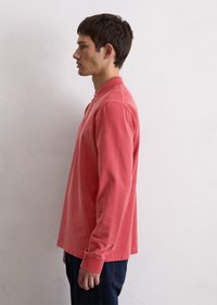 Langermet polo-skjorte i korallfarge, laget av mykt stoff. Har krage og rett snitt, brukt med mørke denim-bukser.