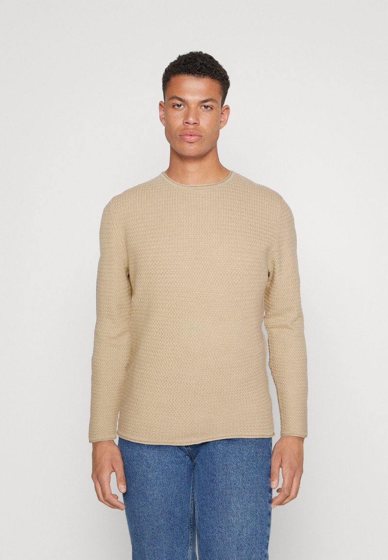 Beige strikket sweater med et tekstureret mønster og rund halsudskæring, parret med blå lige ben jeans.
