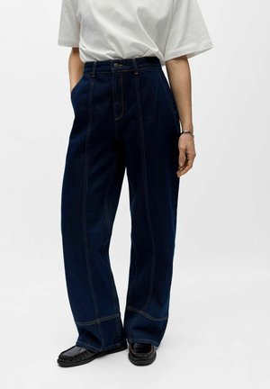 Person trägt dunkelblaue Jeans mit weitem Bein, weißes kurzärmeliges Hemd, schwarze Mokassin-Schuhe und ein silbernes Armband am rechten Handgelenk.