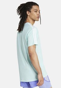 Giovane con lunghe treccine che indossa una maglietta Adidas azzurro chiaro con strisce bianche e pantaloni viola, di profilo contro uno sfondo bianco.