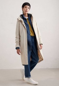 Beige lange jas met een capuchon, gelaagd over een marineblauwe spijkerhemden en een mosterdkleurige trui, gecombineerd met blauwe jeans en witte sneakers.