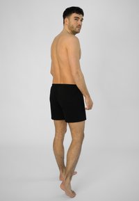 Černé bavlněné šortky s elastickým pasem, předváděné mužem s odkrytou zádí, stojícím na neutrálním pozadí.