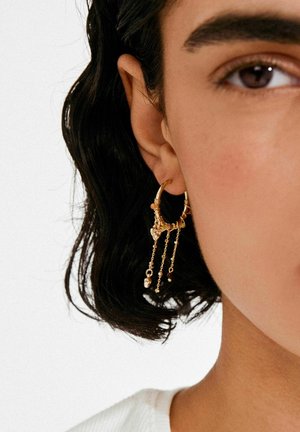 Boucle d'oreille en forme de créole dorée avec des chaînes pendantes et des accents en forme de cœur. Présente un design texturé et des longueurs de chaînes variées pour plus de détails.