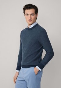 Mornariće plavi pleteni pulover s okruglim izrezom i rebrastim manžetama, složen preko svijetloplavog prugastog košulje, uparen s svijetloplavim hlačama.