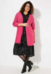 Cardigan rosa a maglia con una vestibilità normale, dotato di maniche testurizzate e scollo aperto, indossato sopra un vestito nero con stampa leopardata. Stivali neri alla caviglia.