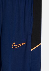 Pantalones deportivos azul marino con un logo de Nike naranja y un panel negro que presenta una franja diagonal naranja en el costado.