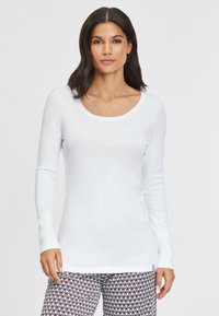 Weißes Langarmoberteil mit rundem Halsausschnitt, aus einem glatten Stoff, kombiniert mit gemusterten Hosen, die ein Zickzackdesign in Rot und Grün aufweisen.