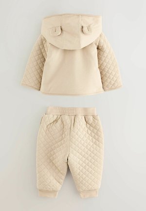 Beige prošívaná mikina s kapucí a odpovídající kalhoty. Obě mají diamantový vzor, měkkou tkaninu a elastické manžety na zápěstí a kotníkách.