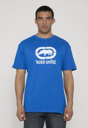 Man wearing a blue T-shirt with white "ecko unltd." text and rhino logo, standing against a plain light gray background.
