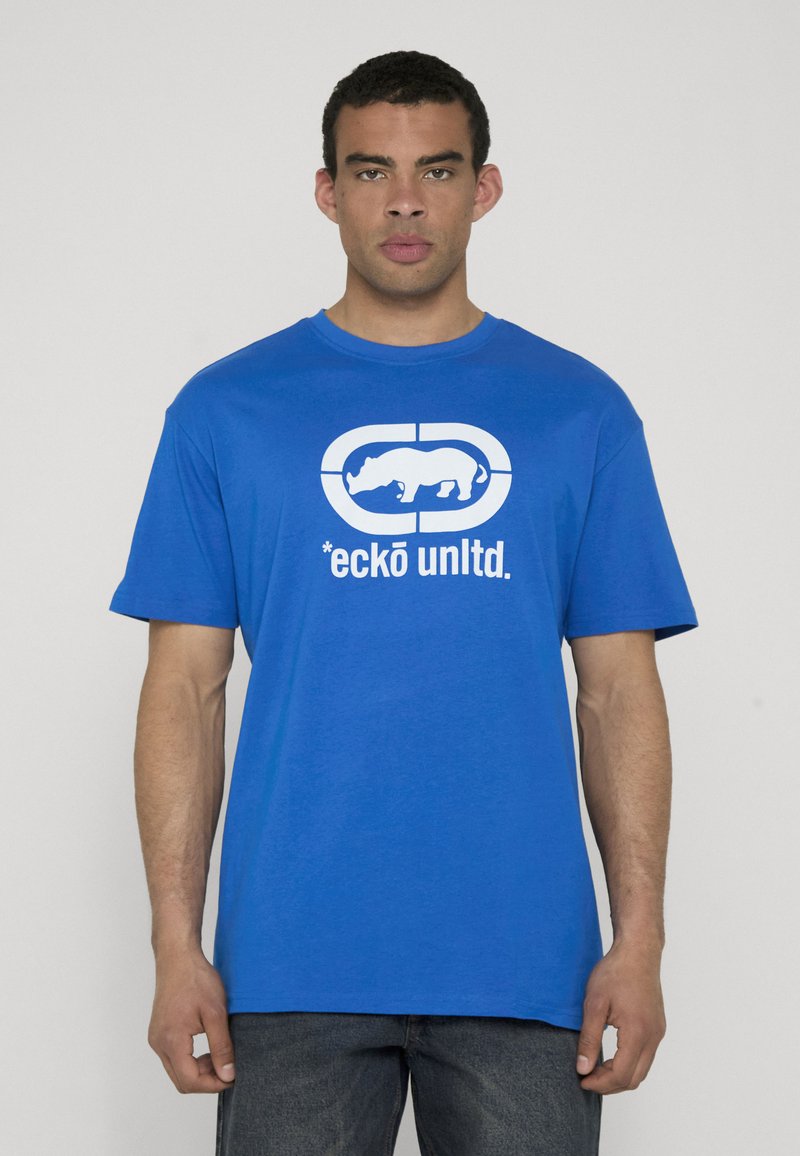 Homme portant un T-shirt bleu avec le texte blanc « ecko unltd. » et un logo rhinocéros, debout devant un fond gris clair uni.