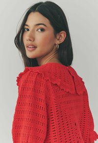 Pull en crochet rouge avec un col en dentelle, présentant un motif de tissage texturé et une fermeture à boutons dans le dos.