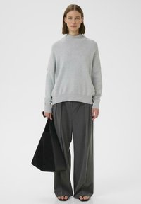 Maglione grigio con collo alto, vestibilità rilassata. Pantaloni ampi grigio scuro, con una borsa tote nera. Sandali aperti completano il look.
