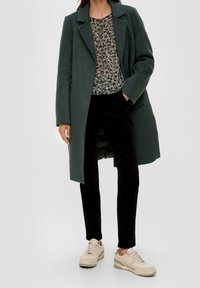 Dunkelgrüner Wollmantel über einem gemusterten schwarz-weißen Top, kombiniert mit schwarzen Samthose und beigen Sneakers mit grünen Akzenten.