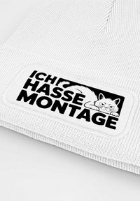 White knitted beanie featuring a rectangular patch with a black cat illustration and bold text reading "ICH HASSE MONTAGE" in black letters.