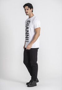 Camiseta blanca con un gran texto negro que dice "GIANNI" y "KANAGH." Llevada con pantalones negros y zapatillas negras, mirando hacia la derecha, contra un fondo liso.