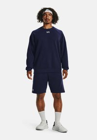 Sudadera azul marino de manga larga con puños de canalé, combinada con pantalones cortos a juego. Lleva una cinta blanca en la cabeza y zapatillas deportivas blancas.