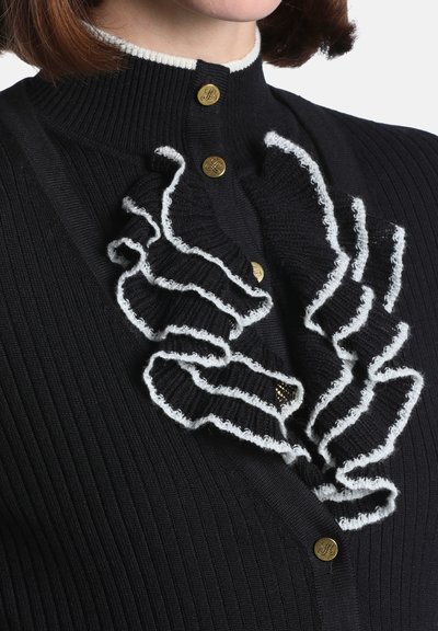 Pull en maille côtelée noire avec des volants bordés de blanc sur le devant et des boutons dorés près du col sur une personne.