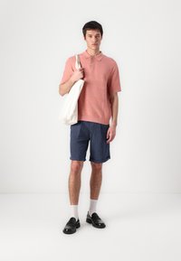 Svijetlo ružičasta polo majica kratkih rukava, teksturirani dizajn; plave traperice kratke nogavice; crne kožne cipele; krema torba; bijele čarape. Minimalistički stil.