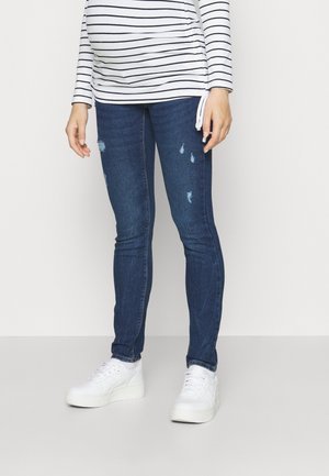 Person iført mørkeblå slidte skinny jeans, hvide platform-sneakers og en hvid langærmet skjorte med sorte vandrette striber.