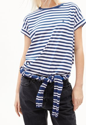 Kvinde iført en hvid og navystribet T-shirt, stoppet ned i sort jeans med et matchende stribet bælte bundet om taljen.