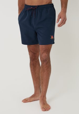 Mężczyzna w granatowych kąpielówkach z logo ośmiornicy w kolorach czerwonym i pomarańczowym na lewej nodze, stojący boso na jednolitym tle.