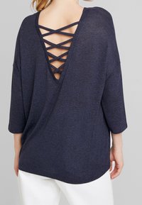 Mörkblå tröja med djup V-rygg och korsade remmar. Tillverkad av texturerad stickad tyg, med tre fjärdedels ärmar och sidslitsar.