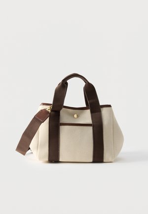 Borsa in tessuto crema con manici marrone scuro, tracolla e bordo, dotata di tasca frontale con dettaglio bottone dorato.